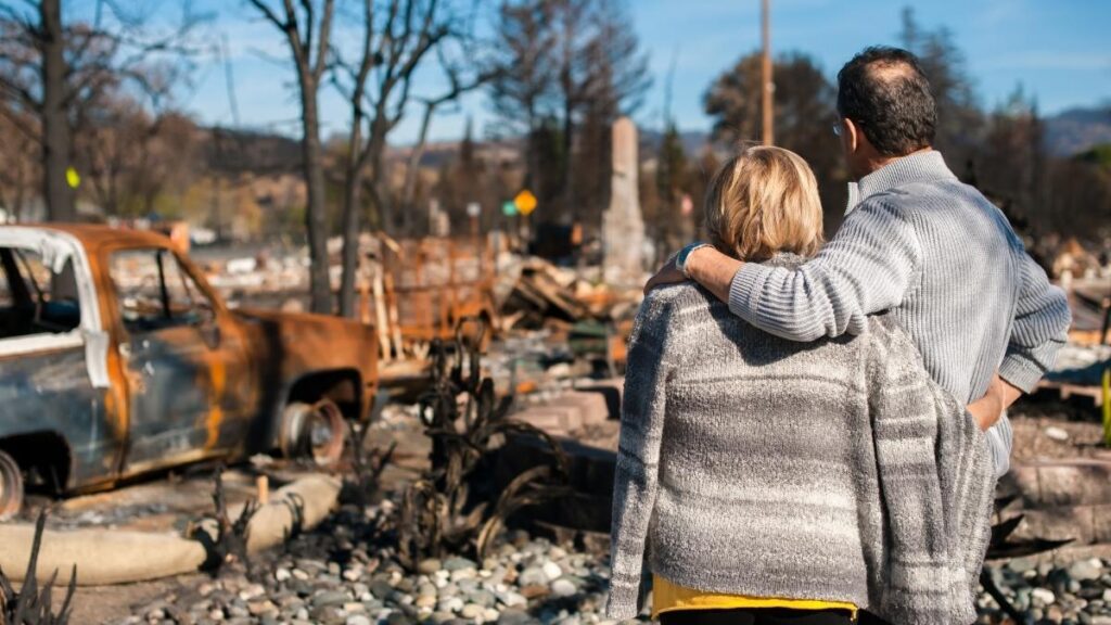 A couple stands together looking at burned vehicles and debris after a wildfire, highlighting the importance of disaster preparedness and recovery planning.