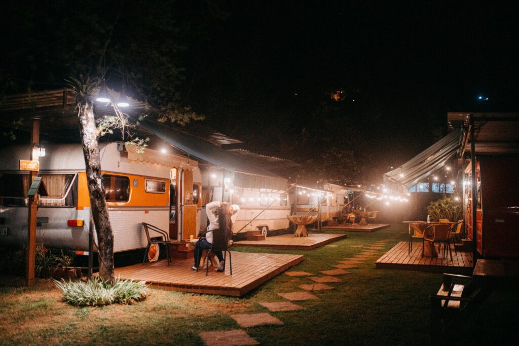 Nighttime view of a cozy RV park with vintage trailers, string lights, and a person relaxing on a small deck.