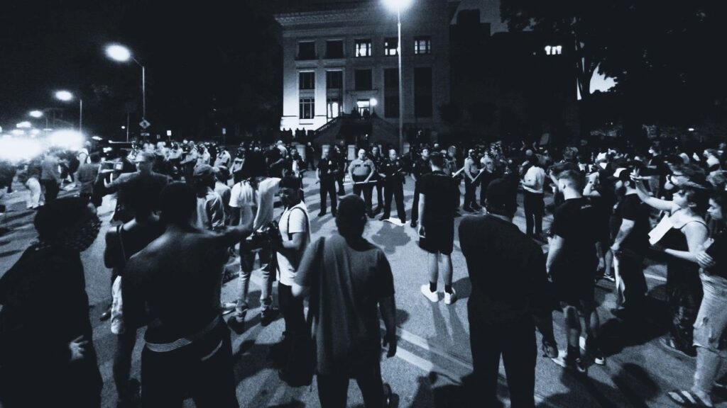 Large crowd of people gathered during a nighttime protest, facing a line of police officers in front of a government building, showing types of disasters to prepare for.