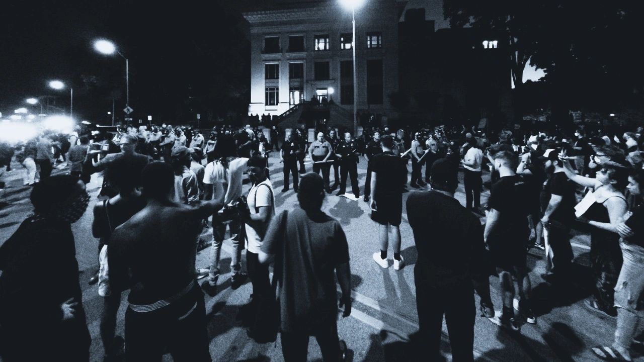 Large crowd of people gathered during a nighttime protest, facing a line of police officers in front of a government building.