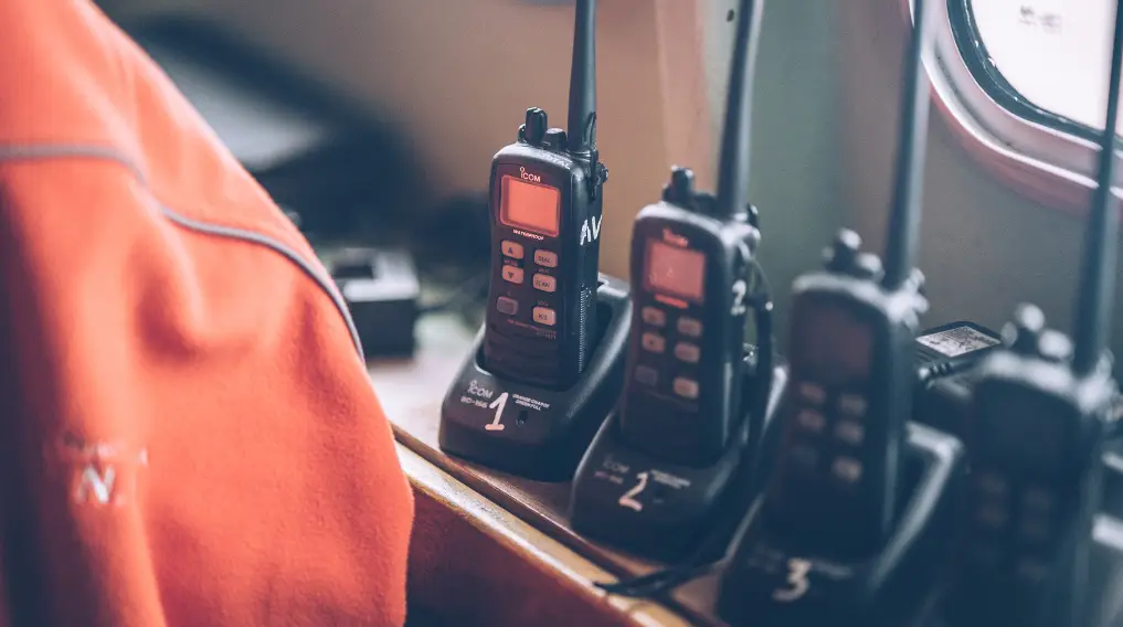 Close-up of handheld emergency radios labeled 1, 2, and 3 charging on a dock near a window and orange jacket.