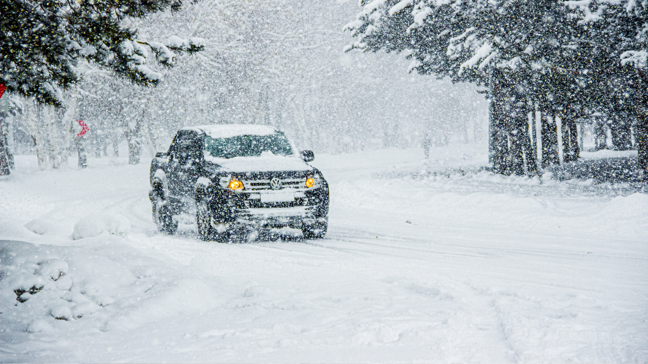 Extreme cold preparedness. SUV driving through heavy snowfall on a snow-covered road surrounded by trees.
