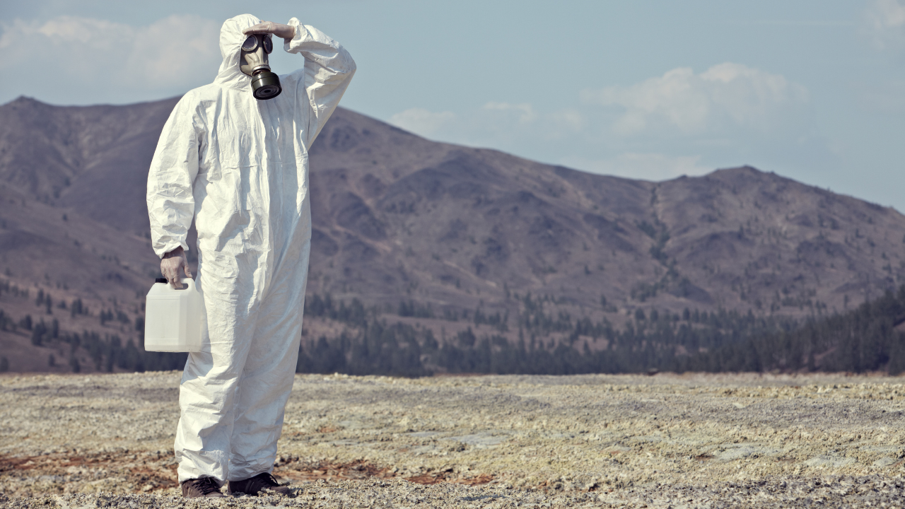 Person in a full hazmat suit and gas mask holding a water container, standing in a barren landscape with mountains in the background.